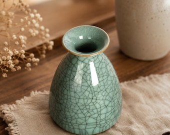 Pode incluir: Um pequeno vaso de cerâmica em forma de sino com um esmalte verde-água claro e um padrão de fissuras escuras proeminente. O vaso apresenta uma borda circular larga e alargada com um acabamento dourado fino, criando uma silhueta suave e redonda.