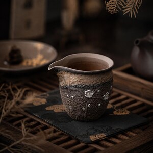 May include: A handcrafted ceramic tea pitcher with a spout, featuring a textured brown and tan exterior. Silver-colored floral accents adorn the dark brown lower section. The pitcher sits on a black mat with gold floral designs, suggesting a tea ceremony setting.