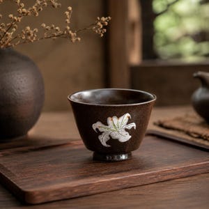 May include: A brown ceramic teacup with a white lily design. The teacup sits on a wooden tray, with a dark vase and dried flowers in the background. The cup has a small base and a flared rim.