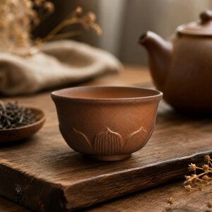May include: A brown ceramic tea cup with a lotus flower design sits on a wooden board. A teapot and a small wooden bowl with tea leaves are in the background. The cup has a rounded shape and a smooth, matte finish.