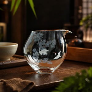 Op de afbeelding: Een heldere glazen theepitcher met een tuit, met een kraanvogel- en dennentakontwerp in wit en grijs. De pitcher staat op een houten oppervlak, met een theekopje op de achtergrond. De kraanvogel heeft een rode snavel.