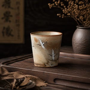 May include: A ceramic cup with a beige and brown glaze, featuring white crane illustrations in flight. The cup has a slightly flared rim and a rustic, handcrafted appearance. The cup is approximately 10 cm tall.