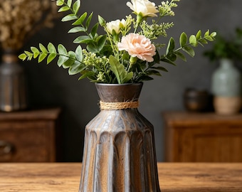 Pode incluir: Um vaso decorativo de cerâmica marrom escuro e dourado, preenchido com um buquê de flores brancas e pêssego e folhagem verde. O vaso tem um design texturizado e canelado e é amarrado com barbante natural ao redor do pescoço. O vaso está em uma mesa de madeira.
