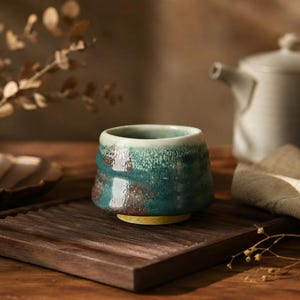 Peut inclure: Une petite tasse à thé en céramique ronde avec un émail réactif aux tons bleu sarcelle profond, bleu et blanc. La surface extérieure présente un aspect texturé et tacheté avec un bord lisse et une base jaune unie distincte. Sans anse.