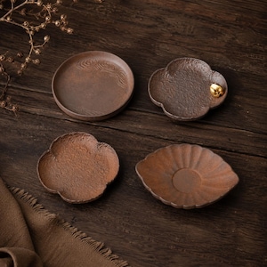 Pode incluir: Quatro pires de chá de cerâmica marrom em várias formas. Um é redondo com um desenho de ondas, dois têm formato de flor e um tem formato de folha. Um pires em forma de flor tem um detalhe dourado. Em uma superfície de madeira escura.