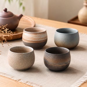 Puede incluir: Cuatro tazas de té de cerámica hechas a mano en tonos tierra variados. Las tazas son redondas con un exterior liso y sin esmaltar. Los colores incluyen tonos de marrón, gris y beige. Una tetera y una bandeja son visibles en el fondo.