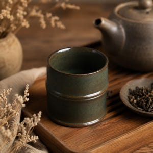 May include: A dark green ceramic tea cup with a bamboo-like design. The cup is on a wooden tray, with a teapot and a small dish of tea leaves in the background. The cup has a smooth, glossy finish.