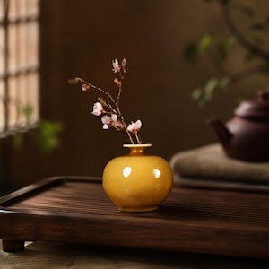 以下が含まれることがあります： 光沢のある黄色の小さな丸い陶器製の花瓶で、首が細くなっています。花瓶には、細い濃い茶色の枝が一本挿されており、開口部から上に向かって伸びる数輪の繊細な淡いピンク色の花が、シンプルでバランスの取れたディスプレイを作り出しています。