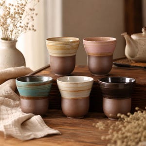 May include: Five ceramic cups with a two-tone design. The bottom half of each cup is a dark brown, while the top halves feature colours like yellow, pink, teal, cream, and dark brown. The cups are arranged on a wooden surface.