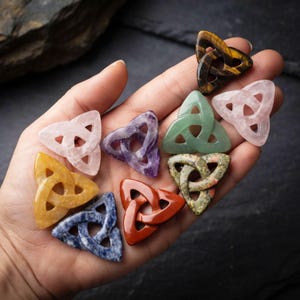 May include: A collection of nine small, triangular-shaped stone charms carved into an interlocking triquetra knot design. The stones feature various colors and textures, including tiger's eye, rose quartz, amethyst, green aventurine, and red jasper.