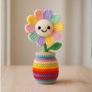 Puede incluir: Una flor de ganchillo hecha a mano en una maceta a rayas de arcoíris. La flor tiene una cara sonriente y pétalos coloridos en tonos de amarillo, rosa, verde, morado y naranja. La maceta también está tejida con una variedad de colores brillantes.