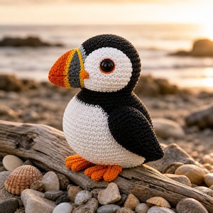 May include: A crocheted puffin toy, featuring a black, white, orange, and grey colour palette. The bird is positioned on driftwood on a beach, with a seashell and pebbles in the foreground. The puffin has orange feet and a large, colourful beak.