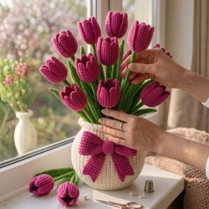 Op de afbeelding: Een handgemaakt gehaakt boeket van roze tulpen in een crèmekleurige vaas met een bijpassende strik. De tulpen hebben groene stelen en bladeren. De vaas staat op een vensterbank.