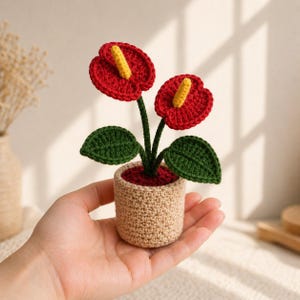 May include: A handmade crochet plant with two red flowers, yellow stamens, and green leaves in a beige pot. The plant is a miniature version of an anthurium, a popular houseplant. The pot is a small, round container.