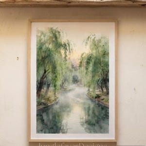 May include: A rectangular framed watercolor painting depicting a river scene with weeping willow trees, hanging on a wall above a wooden table. A woven basket filled with green and red pears sits on the table. The text JunglefeverDesigns is visible.