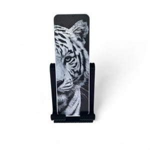May include: A rectangular bookmark featuring a detailed black and white stippled illustration of a tiger's face, held upright in a black plastic stand with a textured base and two vertical supports that cradle the sides of the bookmark.