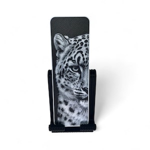 May include: A rectangular black bookmark featuring a detailed white illustration of the right half of a leopard's face with visible spots and whiskers, held upright in a black plastic stand with a textured, ribbed base design.