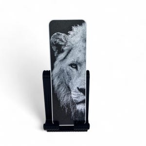 May include: A rectangular bookmark featuring a black and white graphic illustration of a lion's face on the front surface. The bookmark is held upright by a black plastic stand with a ribbed base and vertical side supports for display.