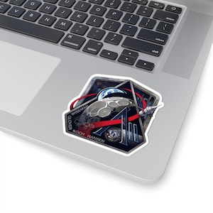 May include: A sticker featuring a space mission patch design. The patch includes a depiction of the moon, Earth, a rocket, and the text "A". The names "WISEMAN", "GLOVER", and "KOCH HANSEN" are also visible.