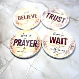 May include: Four round coasters with inspirational messages. The coasters feature phrases like "Believe," "Trust," "Prayer," and "Wait." Each coaster has a floral design and a cross. The text is in a mix of colors, including burgundy and gold.