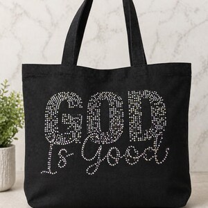May include: Black tote bag with the phrase "GOD is Good" spelled out in sparkling rhinestones. The bag has two black handles and is made of a sturdy fabric. A small potted plant is visible in the background.