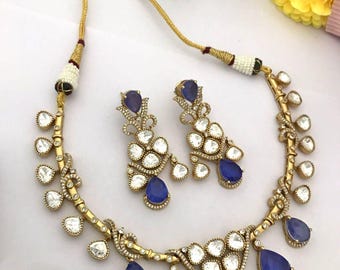 May include: A gold-toned necklace and matching earring set featuring teardrop-shaped blue stones and clear white stones. The necklace has a gold cord with white and black bead accents, while the earrings mirror the teardrop and floral design.