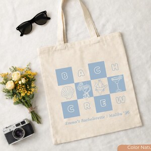May include: A natural canvas tote bag with the words "Beach Crew" in blue squares. The bag features seashell, cocktail, and bikini graphics. Text at the bottom reads "Emma's Bachelorette | Malibu '26". Black sunglasses, a bouquet, and a camera are next to the bag.