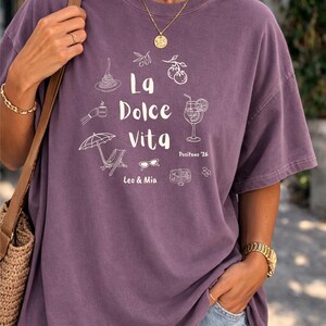 May include: A purple t-shirt with the words "La Dolce Vita" and related illustrations in white. The design includes images of food, drinks, and leisure items. The shirt is a solid color and has a relaxed fit.