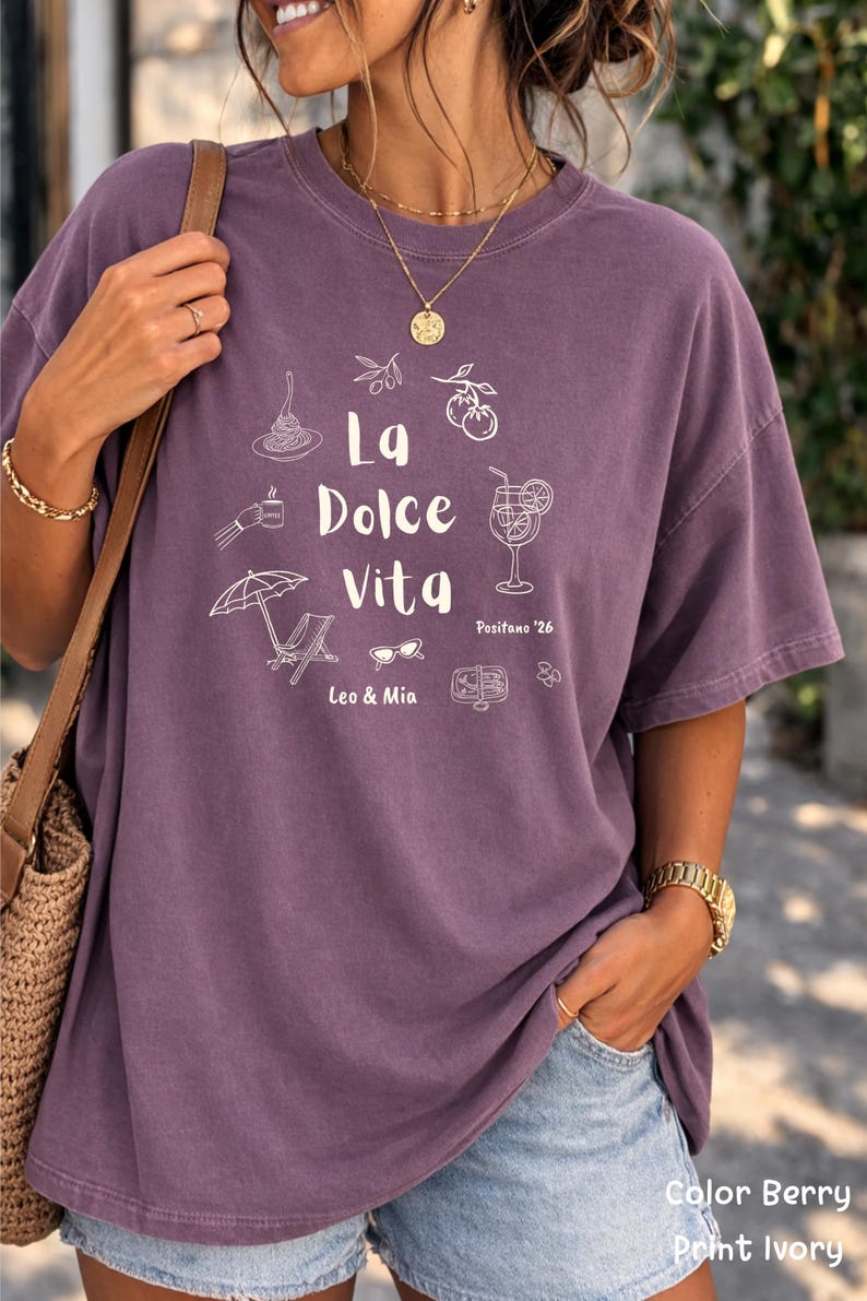 May include: A purple t-shirt with the words "La Dolce Vita" and related illustrations in white. The design includes images of food, drinks, and leisure items. The shirt is a solid color and has a relaxed fit.
