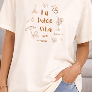 May include: Ivory-colored t-shirt with a terracotta print. The design includes the words "La Dolce Vita" with illustrations of pasta, a cocktail, sunglasses, and a beach umbrella. The text "Positano '26" and "Leo & Mia" are also included.