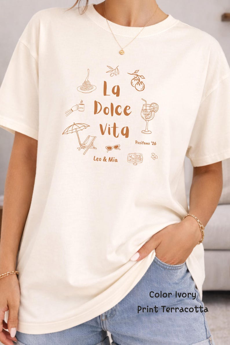 May include: Ivory-colored t-shirt with a terracotta print. The design includes the words "La Dolce Vita" with illustrations of pasta, a cocktail, sunglasses, and a beach umbrella. The text "Positano '26" and "Leo & Mia" are also included.