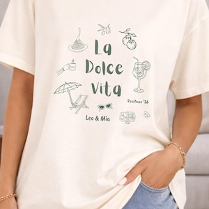 May include: Ivory-colored t-shirt with a green graphic design. The design includes the text "La Dolce Vita" and illustrations of food, drinks, and leisure items. The text "Positano '26" and "Leo & Mia" are also included.