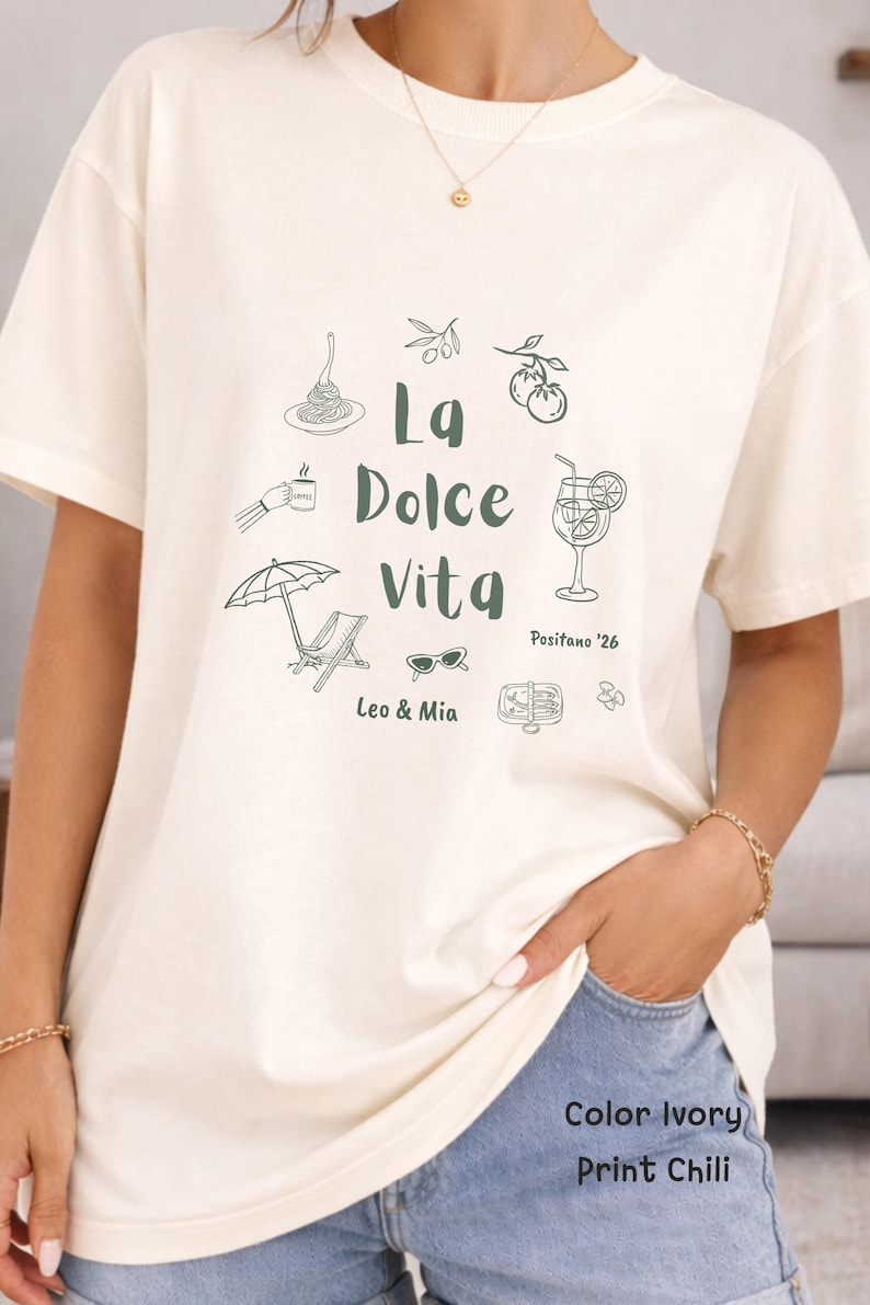 May include: Ivory-colored t-shirt with a green graphic design. The design includes the text "La Dolce Vita" and illustrations of food, drinks, and leisure items. The text "Positano '26" and "Leo & Mia" are also included.