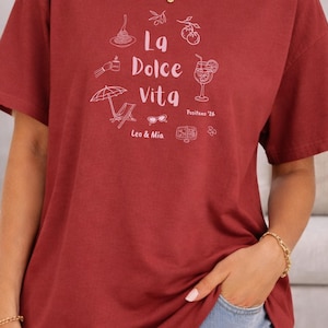 May include: A maroon t-shirt with a white graphic design featuring the words "La Dolce Vita" and illustrations of Italian food and leisure items. The shirt also includes the text "Positano '26" and "Leo & Mia".