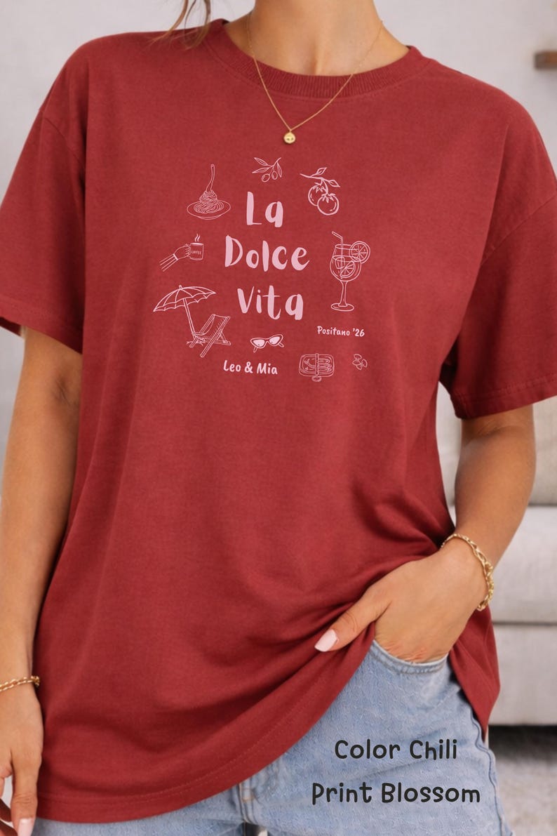 May include: A maroon t-shirt with a white graphic design featuring the words "La Dolce Vita" and illustrations of Italian food and leisure items. The shirt also includes the text "Positano '26" and "Leo & Mia".