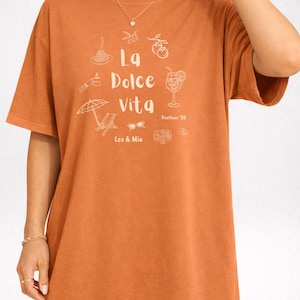 May include: A short-sleeved, orange t-shirt with the phrase "La Dolce Vita" and related illustrations in white. The shirt also features the text "Positano '26", "Leo & Mia", and the color and print details.