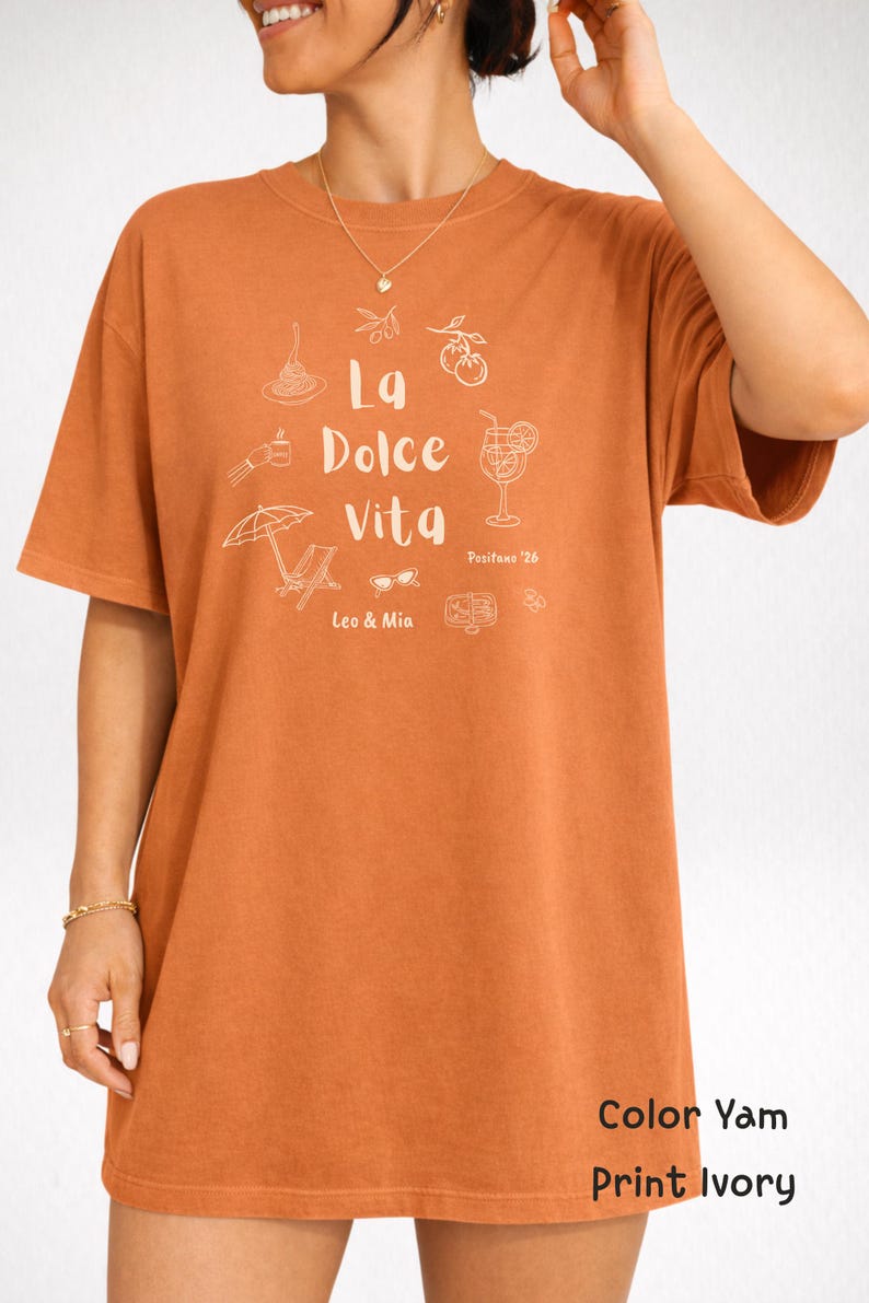 May include: A short-sleeved, orange t-shirt with the phrase "La Dolce Vita" and related illustrations in white. The shirt also features the text "Positano '26", "Leo & Mia", and the color and print details.