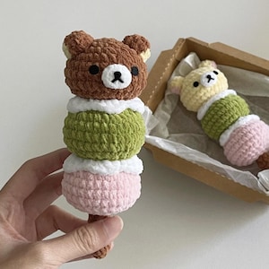 May include: Crocheted bear-shaped dango, a Japanese sweet treat, in brown, green, and pink. The dango is held by a hand, with another in a box. The bear has a black nose and eyes. The dango has a white layer between the colors.