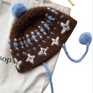 May include: A brown knit hat with white star and blue accents. The hat has a blue pom-pom on top and two blue dangling strings with pom-poms. The hat is resting on a white tote bag.