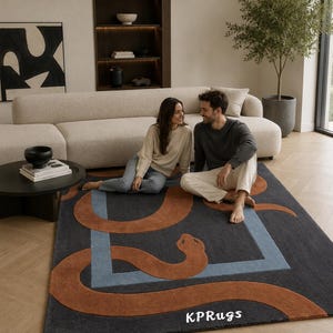 May include: A rectangular area rug featuring a dark gray background with a large, winding rust-colored snake motif and a light blue geometric square frame. The text KPRugs is printed in white in the bottom right corner of the rug surface.
