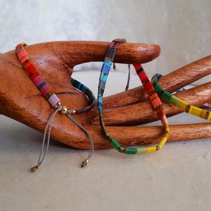 Puede incluir: Cuatro pulseras coloridas hechas de pequeñas baldosas cuadradas con un cordón gris y cuentas doradas. Las pulseras están dispuestas en una mano de madera.