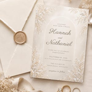 Può includere: Un invito di nozze color crema con un design ad arco, accenti floreali dorati e il testo Hannah and Nathaniel, accompagnato da una busta abbinata sigillata con ceralacca dorata e due anelli dorati su una superficie strutturata.