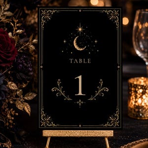 May include: A rectangular black table number card featuring the text TABLE 1 in gold. The design includes a gold crescent moon, stars, and ornate filigree corner accents. The card is displayed on a small, textured gold metal easel stand.
