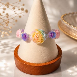 Könnte beinhalten: Ein farbenfrohes Armband mit blütenförmigen Perlen in Rosa, Lila, Gelb und Blau. Das Armband wird auf einem cremefarbenen, kegelförmigen Ständer mit Holzsockel präsentiert, der sein zartes Design und seine leuchtenden Farben hervorhebt.