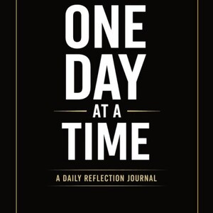 May include: Black journal cover with the words "ONE DAY AT A TIME" in large white letters. Below the text is "A DAILY REFLECTION JOURNAL" in smaller white letters. The journal has a gold border.