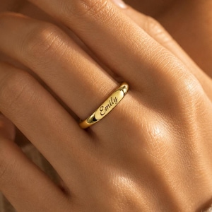 May include: A polished gold-tone band ring featuring a smooth, rounded surface with the name Emily engraved in a cursive script font. The ring has a simple, continuous circular design with a reflective metallic finish and a centered inscription.