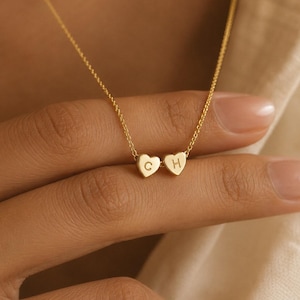 May include: A gold-toned necklace featuring two small heart-shaped charms suspended from a delicate chain. Each heart charm is engraved with a single uppercase letter, specifically the letters C and H, centred on the front of each heart.