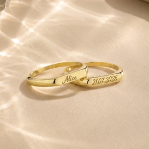 Puede incluir: Dos anillos de tono dorado con un acabado liso y pulido. Un anillo está grabado con el nombre Alice en letra cursiva, y el otro anillo está grabado con la fecha 21.07.2026 en una fuente sans-serif clara. Ambos anillos tienen un diseño plano.