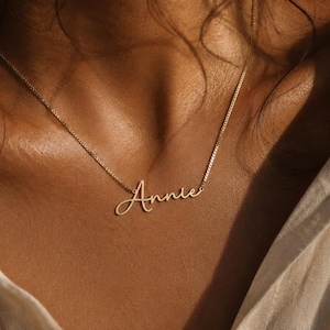 Puede incluir: Un collar con placa de metal en tono dorado que presenta el nombre Annie escrito en una fuente cursiva. El colgante está unido a una delicada cadena de eslabones cuadrados que descansa sobre la clavícula, reflejando luz cálida.