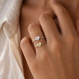 Op de afbeelding: Twee dunne metalen ringen gedragen aan een vinger. De bovenste ring is zilverkleurig met een klein hartvormig bedeltje met de letter H. De onderste ring is goudkleurig met een klein hartvormig bedeltje met de letter A. Beide ringen hebben een eenvoudige band.